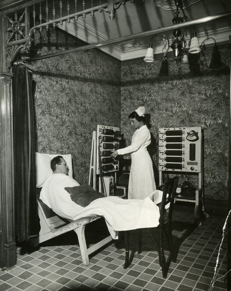 Photographie en noir et blanc représentant une petite pièce sans fenêtre. Le sol est recouvert d’un vinyle à motif carrelage et les murs sont recouverts de papiers peints aux motifs botaniques. Un homme, le patient, est couché sur une sorte de chaise longue. Une femme en habit d’infirmière manipule des machines qui ressemblent à des tableaux sur pattes avec des boitiers noirs installés dessus.