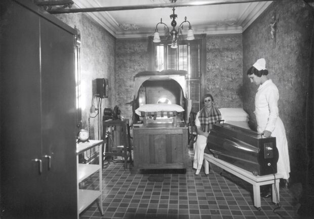 Photographie en noir et blanc représentant une petite pièce avec une fenêtre à volets fermés. Il y a un luminaire au plafond et un crucifix au mur. Quelques étagères sont installées et portent de petits appareils impossibles à identifier. Une sorte de lit qui ressemble à une grande boîte en bois avec un oreiller se trouve au centre. Une femme est debout en habit d’infirmière et pointe une machine ressemblant à un canon foncé vers une personne qui semble être une patiente de l’Institut.