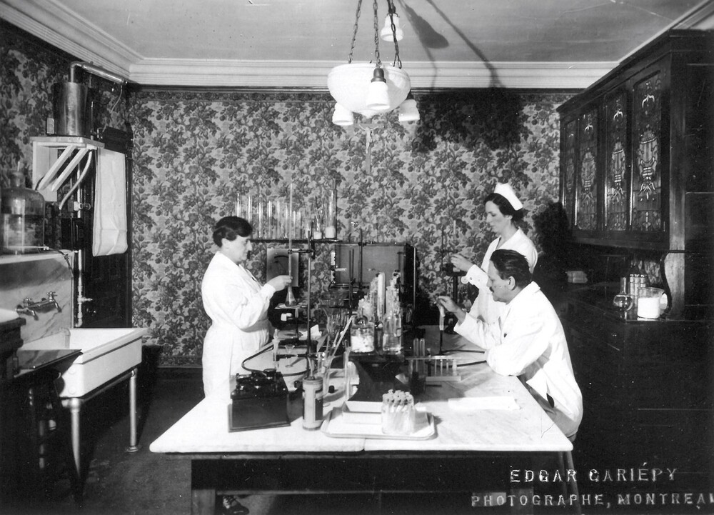 Photographie en noir et blanc d’une salle sans fenêtre dont les murs sont tapissés de papier-peint fleuri. Une lampe est suspendue au plafond, une grande armoire est sur le mur de droite et un grand lavabo sur le mur de gauche. Au centre se trouve une large table sur laquelle est installée une panoplie d’instruments de laboratoires. Éprouvettes, béchers et autres instruments moins reconnaissables sont manipulés par deux femmes en habit d’infirmière et un homme portant un sarrau. 