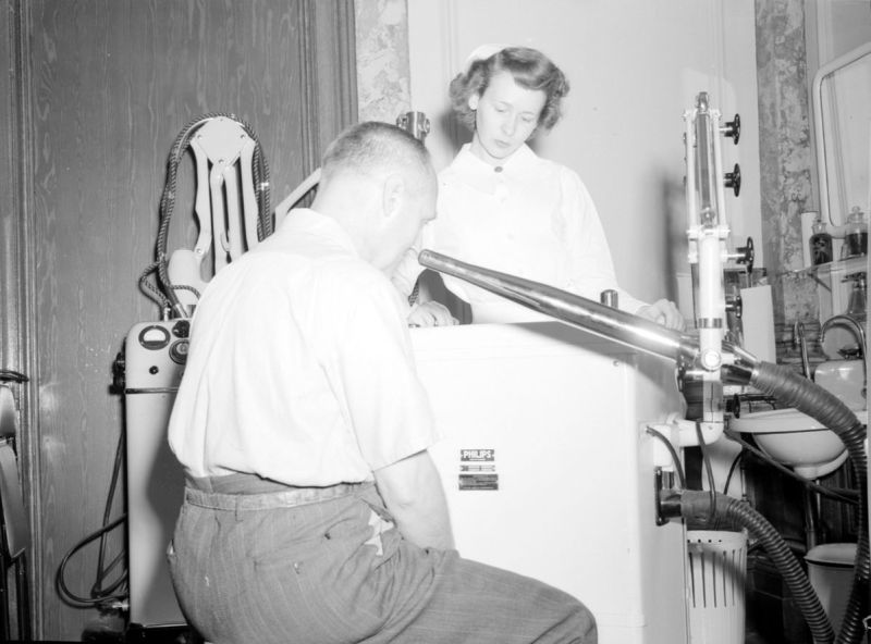 Photographie en noir et blanc sur laquelle se trouve une femme debout en habit d’infirmière en second plan et un homme assis en premier plan. Une machine qui a une forme de boîte les sépare. Une sorte de tuyau sort de la machine et est pointé au visage de l’homme assis. 