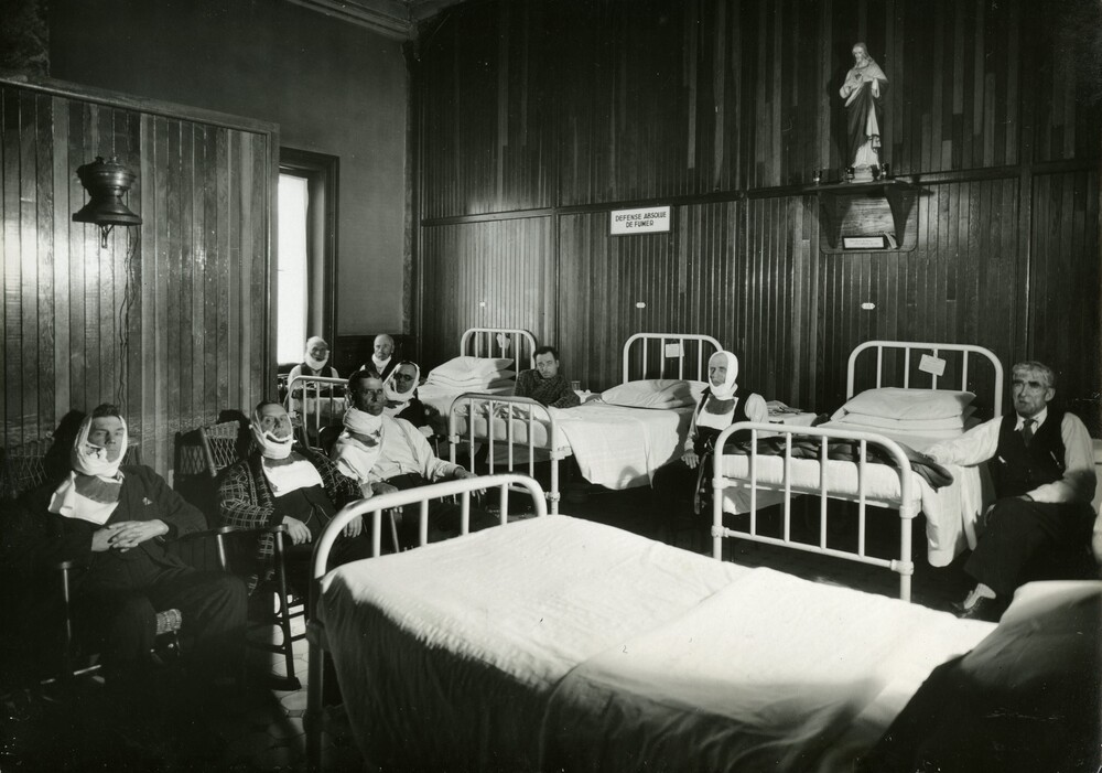 Photographie en noir et blanc représentant une salle meublée de lits d’hôpitaux et de chaise. Neuf hommes sont assis, la plupart ayant des bandages autour du cou ou du visage. Une petite statue à l’effigie de Jésus trône sur une étagère et un panneau sur lequel est écrit «Défense absolue de fumer» est installé sur le même mur.