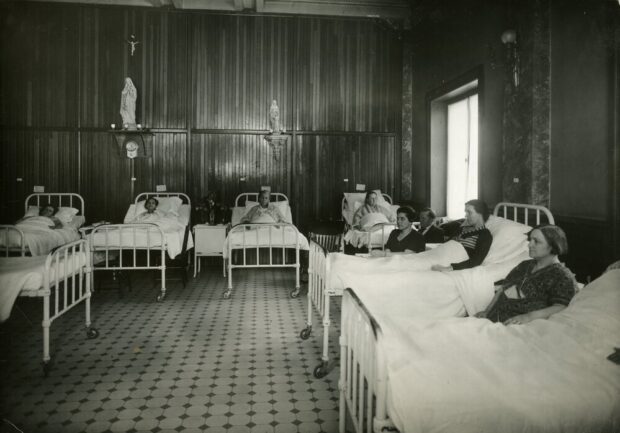 Photographie en noir et blanc représentant une salle meublée de lits d’hôpitaux et de chaise. Deux femmes sont assises et 7 autres sont couchées dans les lits. Deux petites statues à l’effigie de Marie, la mère de Jésus, trônent sur des étagères. Un petit crucifix est installé au-dessus d’elles.