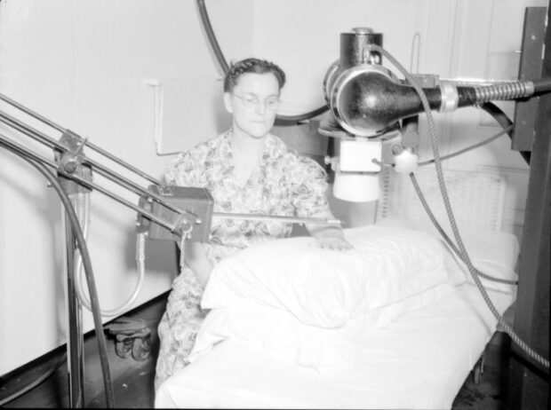 Photographie en noir et blanc représentant une femme assise mettant sa main sur un oreiller placé sur un lit d’hôpital. Deux machines sont installées sur des supports autour d’elle. L’une est une tige de métal tenue à l’horizontale par dessus sa main et l’autre ressemble à une caméra dont on pointe l’objectif par dessus sa main. 