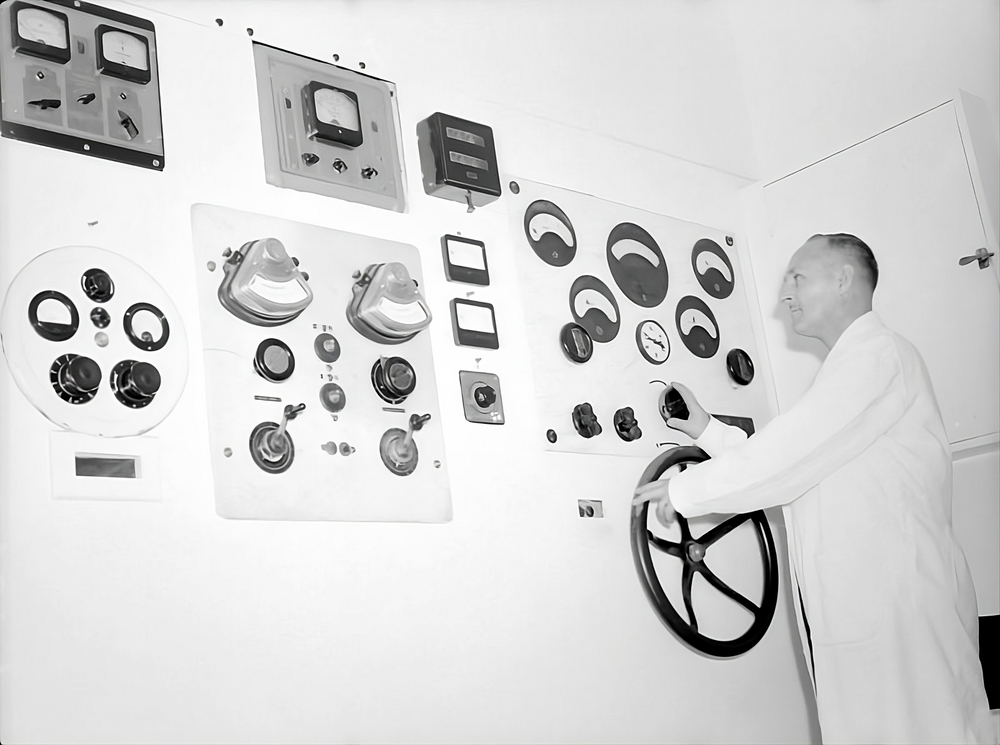 Photographie en noir en blanc d’un homme à droite de l’image en train d’utiliser une grande machine qui prend tout le reste de l’espace de l’image. La machine est un grand mur blanc sur lequel différent cadrans et manivelle sont posées. 