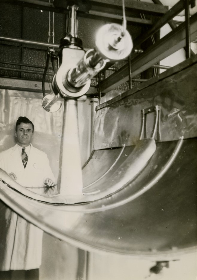 Photographie en noir et blanc d’une machine en premier plan et d’un homme en sareau derrière. La machine est suspendue et ressemble à une sorte de petit canon de forme cylindrique.