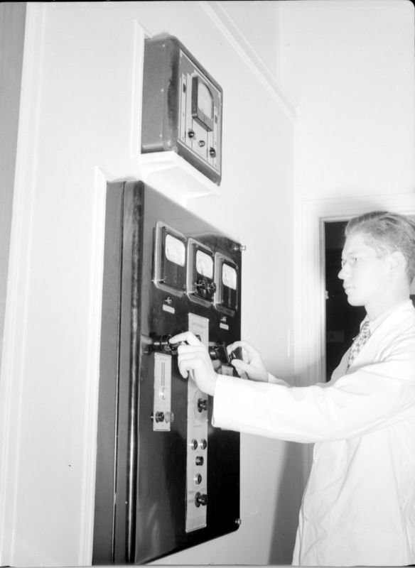 Photographie en noir et blanc d’un jeune homme debout de côté habillé d’un sarreau. Il manipule des boutons rotatifs sur une machine encastrée dans un mur blanc. La machine est constituée de ces boutons ainsi que plusieurs cadrans à aiguilles.