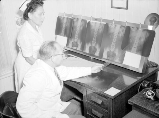 Photographie en noir et blanc représentant une femme et un homme dans une petite salle. L’homme, assis dans une chaise à un bureau, porte un sarrau et pointe avec une règle un tableau lumineux sur lequel des radiographies de plastique transparent sont épinglées. La femme debout est en habit d’infirmière et l’assiste. 