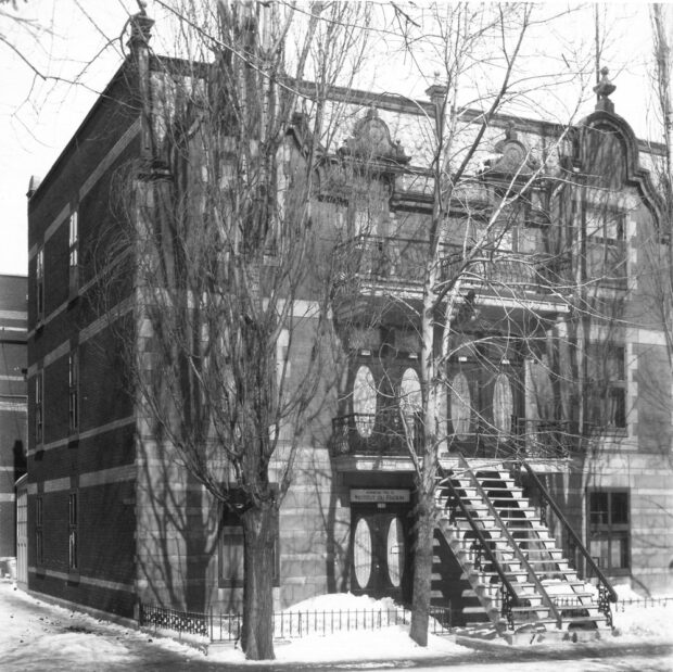 Photographie en noir et blanc représentant la façade d’un imposant triplex partiellement caché derrières des arbres. De la neige recouvre le sol. Le bâtiment de huit logements possède aussi deux balcons avants.