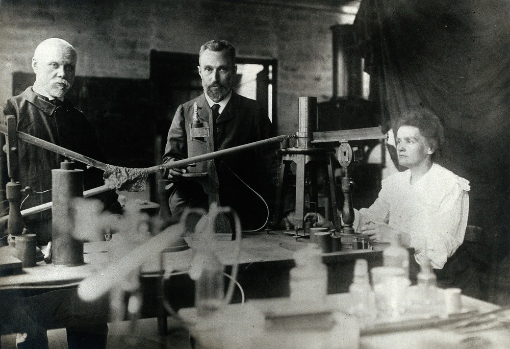 Photographie en noir et blanc représentant un laboratoire avec plusieurs instrument scientifiques sur une table. 3 personnes sont installées à la table. Deux hommes sont debout face à la caméra à gauche et une femme est assise en train de manipuler des instruments de laboratoire à gauche. 