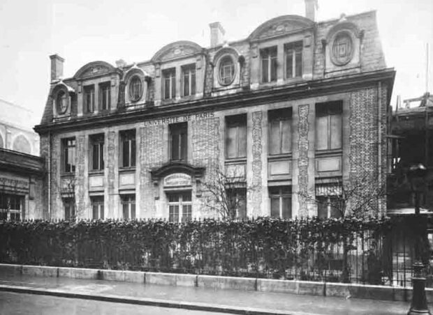 Photographie en noir et blanc représentant la façade d’un bâtiment de trois étages en brique avec d’imposantes lucarnes de fenêtres au troisième étage. Au-dessus de l’entrée il est écrit «Université de Paris». Le bâtiment est entouré d’une clôture en métal à travers laquelle pousse une vigne. 