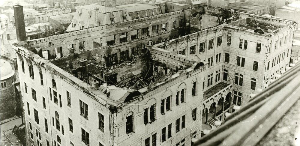 Photographie en noir et blanc, prise en plongé, d’un grand bâtiment de cinq étages dont le plus haut est complètement détruit à la suite d’un incendie. 