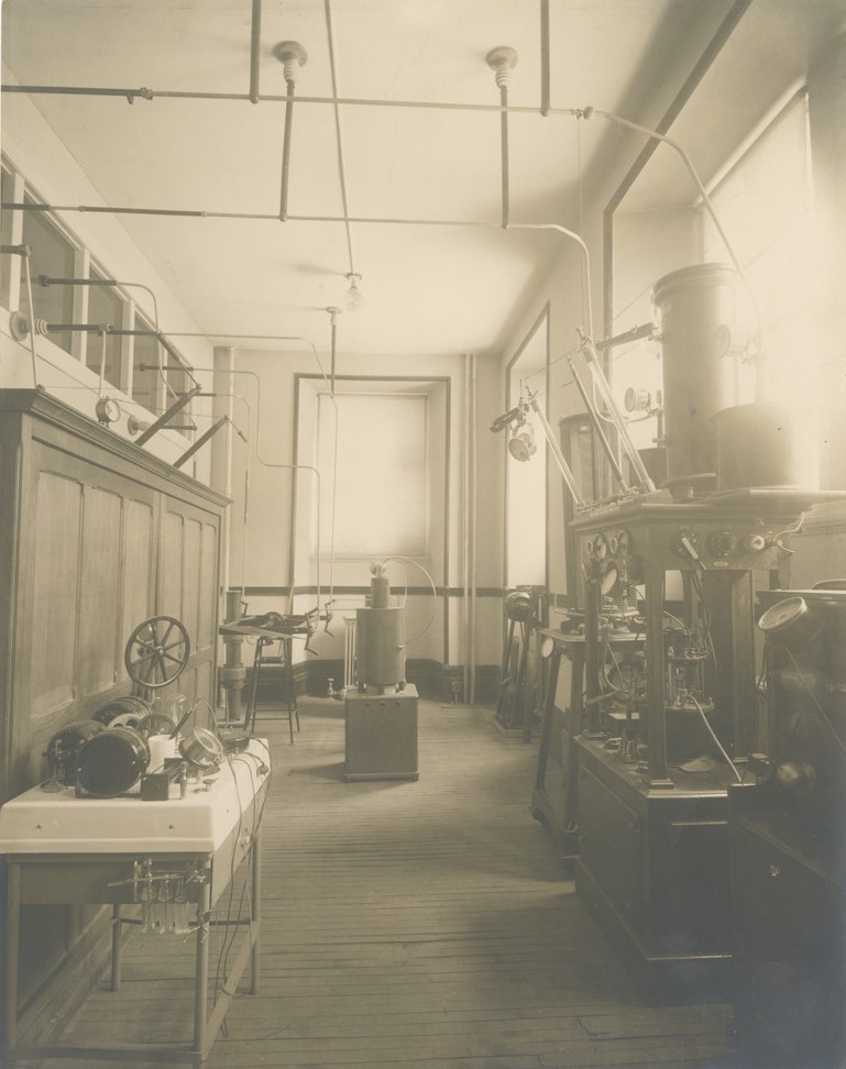 Photographie en noir et blanc d’une petite salle construite sur le long aux murs blancs et au plancher de lattes de bois. Elle est remplie de différentes machines construites de rouages, de récipients de verres, de cadrans et de tubes. 