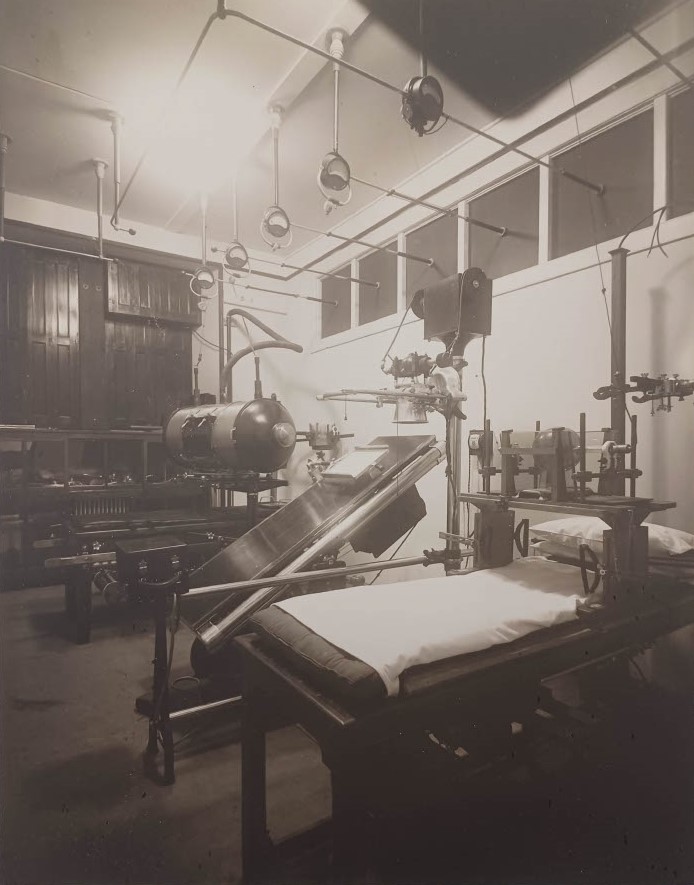 Photographie en noir et blanc représentant une pièce remplie d’appareils et de tables aménagés en lits d’hôpital. Les appareils sont variés, certains ressemblant à des espèces de barils, d'autres à d’anciennes caméras photographiques.  