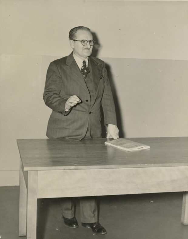 Photographie en noir et blanc représentant un homme debout derrière une table de bois portant un trois-pièces. L’homme porte des lunettes et semble gesticuler quelque chose. Une revue quelconque est le seul élément posé sur la table.