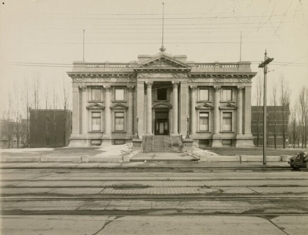 Photographie en noir et blanc d’un imposant bâtiment de style beaux-arts avec un chemin bétonné menant vers l’escalier et les portes centrales. À l’avant-plan, se trouve la rue pavée de briques.