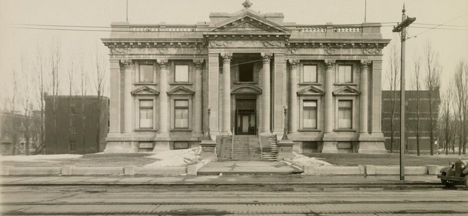 Photographie en noir et blanc d’un imposant bâtiment de style beaux-arts avec un chemin bétonné menant vers l’escalier et les portes centrales. À l’avant-plan, se trouve la rue pavée de briques.