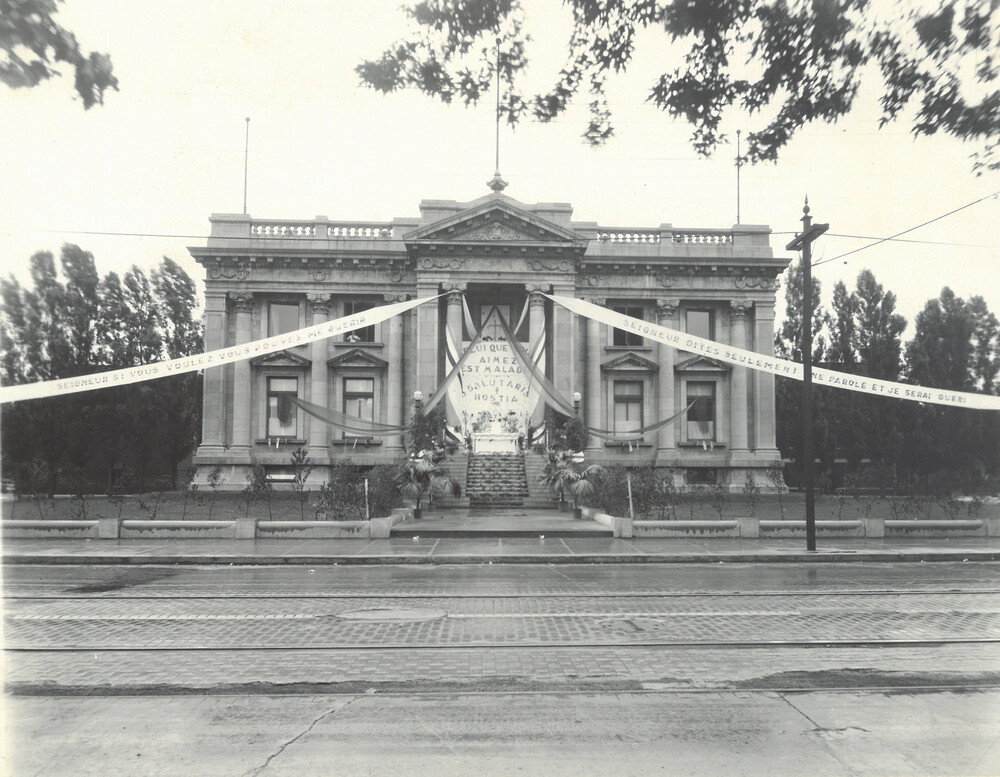 Photographie en noir et blanc d’une grand batiment style beaux-arts. Sa façade est décorée de banderoles attachées à ses colonnes. Sur les banderoles il est écrit : «Seigneur si vous voulez vous pouvez me guérir. Seigneur dites seulement une parole et je serai guéri. Celui que vous aimez est malade. Salutaris hostia.» 