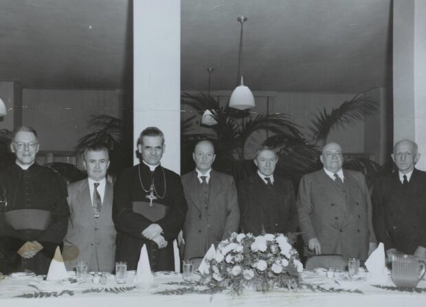 Photographie en noir et blanc représentant sept hommes debout dans un grand hall derrière une table de banquet. La plupart d’entre eux sont habillés en trois pièces mais deux d’entre eux portent la soutane. 