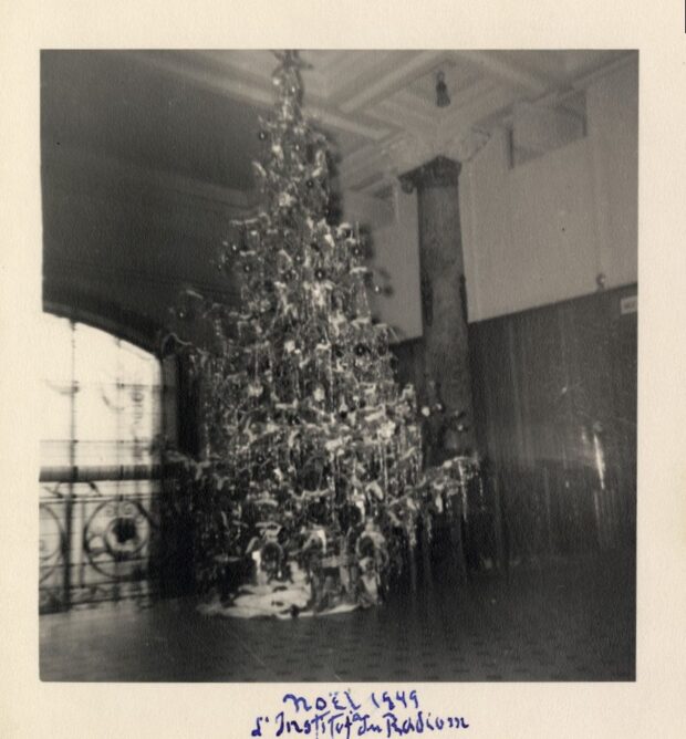 Photographie en noir et blanc et un peu floue d’un sapin richement décoré situé dans un hall.