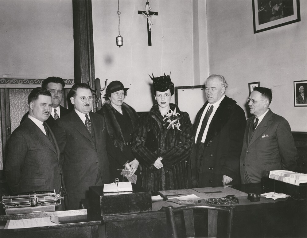 Photographie en noir et blanc représentant sept personnes bien habillées derrière un bureau. Il y a cinq hommes et deux femmes au centre. Le groupe se trouve dans une pièce aux plafonds hauts comportant une fenêtre ainsi que des photos et un crucifix accrochés aux murs. Les seuls meubles sont un bureau et une chaise en bois.