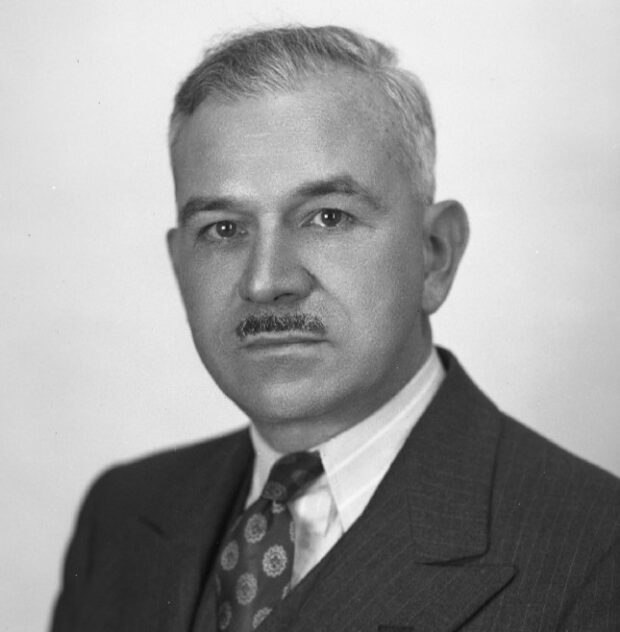 Photographie en noir et blanc représentant un homme de face en veston cravate. Il a les cheveux courts grisonnants et une petite moustache.