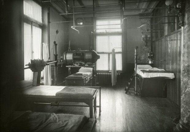 Photographie en noir et blanc d’une salle au plafond haut bien éclairée par deux grandes fenêtres tamisée par des rideau mince. Trois lits sont bien visibles sur l’image et disposés de manière à pouvoir accueillir des installations de machines par dessus. Les trois machines ressemblent à des boîtes en métal et l’une d’elles à un grand tonneau couché sur la longueur. 