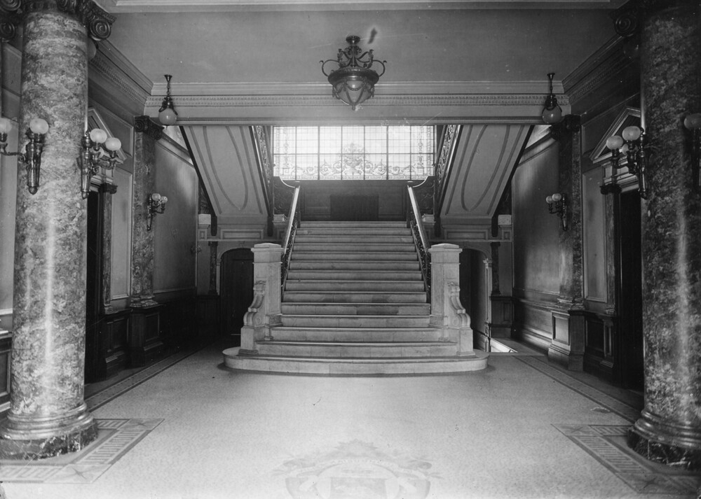 Photographie en noir et blanc d’un hall d’entrée menant vers un escalier. Le hall a des allures beaux-arts avec des piliers de marbre et des murs blancs. Des lumières sont attachées aux piliers et suspendues au plafond. Sur le plancher, on peut voir en carrelage des armoiries très pâles. 