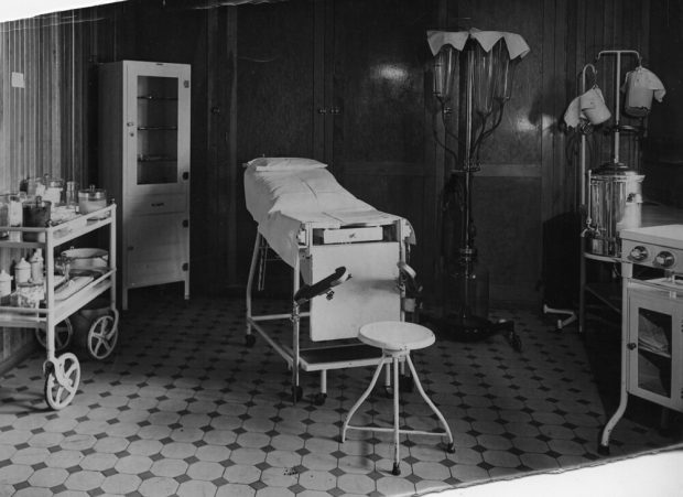 Photographie en noir et blanc d’une salle aux murs de bois laqués et au plancher de carrelage. La salle est meublée d’un lit d'hôpital, d’armoires, d’une table à roues pleine de matériel médical et de quelques machines constituées de grandes cuves de verre et d’acier. 