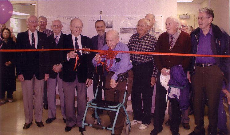 Un groupe de 12 hommes lors d'une cérémonie d'inauguration. Le ruban est rouge, avec un grand nœud au centre. En chemise bleue, un homme avec marchette prend les ciseaux de la main droite pour couper le ruban.