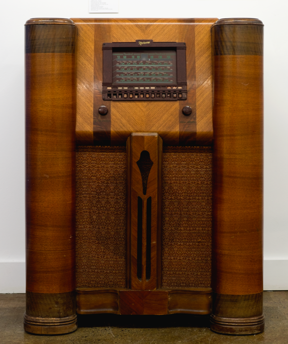 La radio se trouve dans un grand meuble fait de bois de différentes couleurs. Au-dessus de l'écran du haut-parleur se trouvent les deux cadrans, l'un pour le volume et l'autre pour régler la fréquence. Plus haut se trouve une fenêtre à cadran carré.