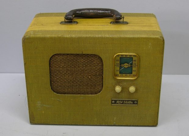 En forme de rectangle avec une poignée sur le dessus, un haut-parleur au milieu, deux cadrans sur le côté, avec un affichage sur le dessus. La radio a une finition en tissu jaune-vert.