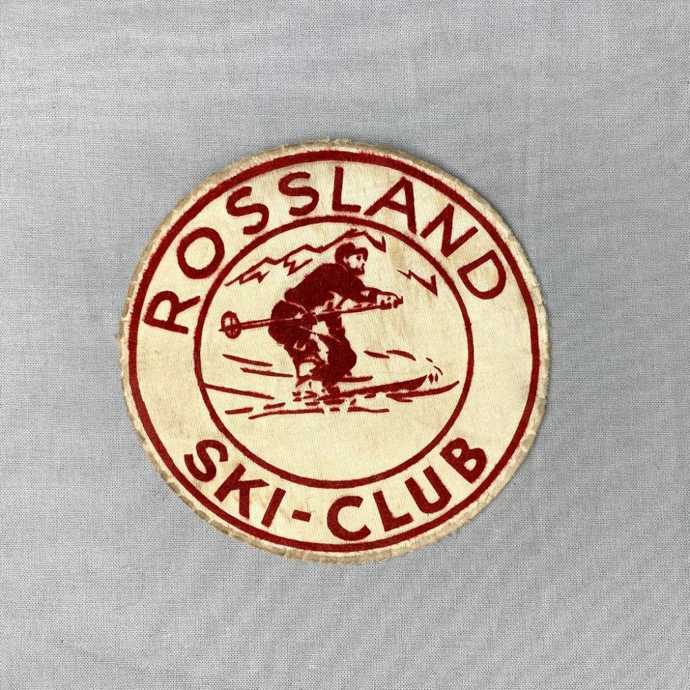 Écusson rond, beige avec une inscription en rouge et une silhouette de skieur au centre.