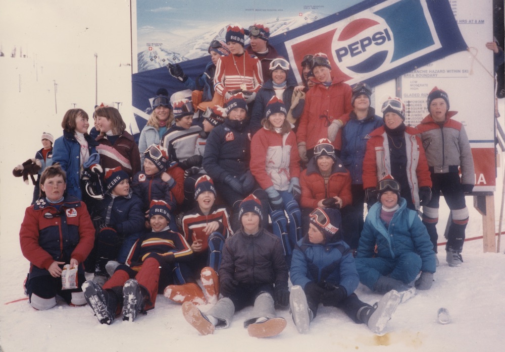 26 enfants en tenue de ski avec des tuques des Red Mountain Racers posent sur une montagne enneigée devant une bannière de commanditaire Pepsi drapée en travers d’un plan des pistes.