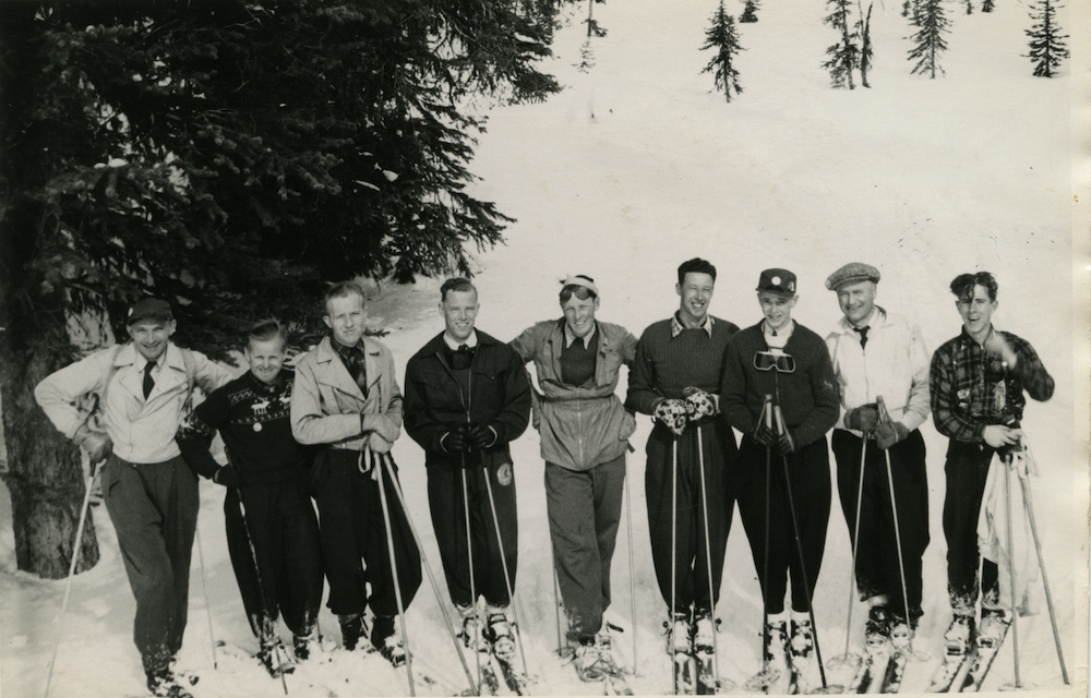 Groupe de neuf hommes sur des skis photographiés sur une montagne enneigée avec des arbres en arrière-plan."