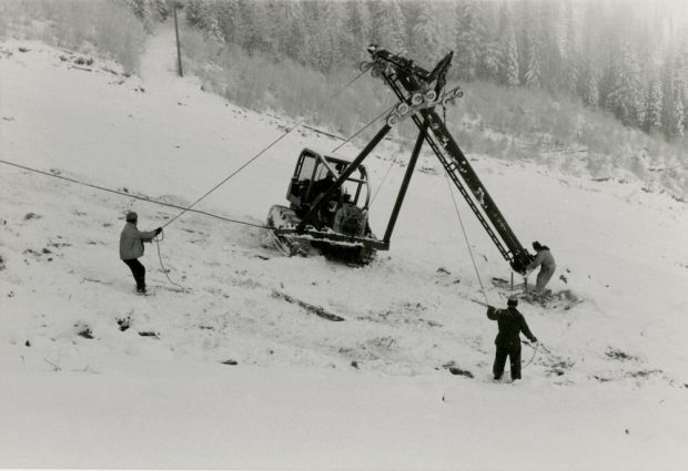 Photo en noir et blanc d’une grue avec trois hommes en train d’installer un pylône sur une pente de ski.