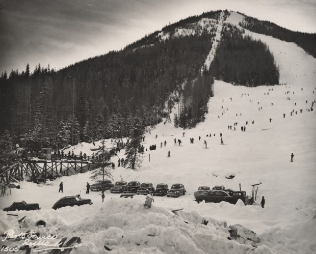 Photo en noir et blanc montrant le télésiège, la piste et le stationnement de Red Mountain. De nombreux skieurs sont en train de descendre ou debout dans la file d’attente du télésiège, on peut distinguer 14 voitures dans le stationnement.