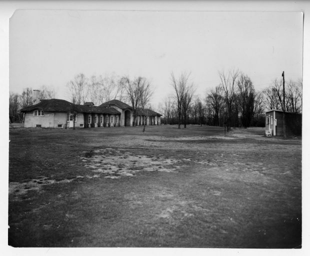 Photographie en noir et blanc du chalet du Drummondville Golf & Country Club. La bâtisse construite sur la longueur est couverte de plantes grimpantes et est entourée d’arbres.