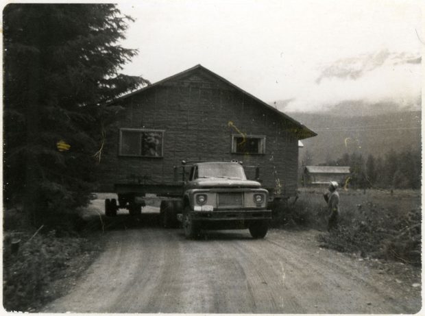 Photographie en noir et blanc montrant un camion plate-forme qui déménage une maison. Le camion est sur une route de terre, bordée d’arbres à gauche, et un homme debout de l’autre côté.