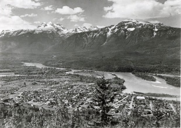 Un glacier à trois pics et d’autres montagnes dominent une petite ville, entre lesquelles coule une rivière. La photographie en noir et blanc a été prise depuis le flanc d’une montagne, offrant une vue plongeante sur la ville en bas.