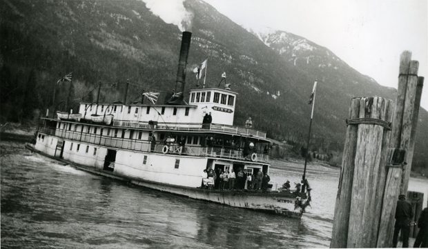 Photographie en noir et blanc. Un bateau à vapeur est sur l'eau avec des montagnes en arrière-plan. Des gens sont à l'avant du navire, plusieurs drapeaux sortent du navire et une grande couronne est accrochée à l'avant du bateau.