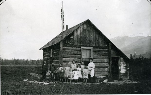 Photographie en noir et blanc. Douze écoliers se tiennent à l’extérieur d’une petite école en bois avec leur enseignante. L’école est dotée d’une porte sur la droite, une fenêtre sur le devant et sur le côté, et un mât de drapeau à l’arrière. Une petite quantité de neige est encore visible au sol autour de l’école. Il y a des arbres et des montagnes aux contours flous en arrière-plan.