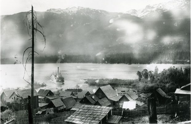 Photographie en noir et blanc. Vue d'une ville, montrant plusieurs maisons, orientée vers un plan d'eau. Un bateau à vapeur s’approche de la ville. Les montagnes sont en arrière-plan. Un arbre mort avec quelques branches bloque légèrement la vue à gauche, dans la photo.