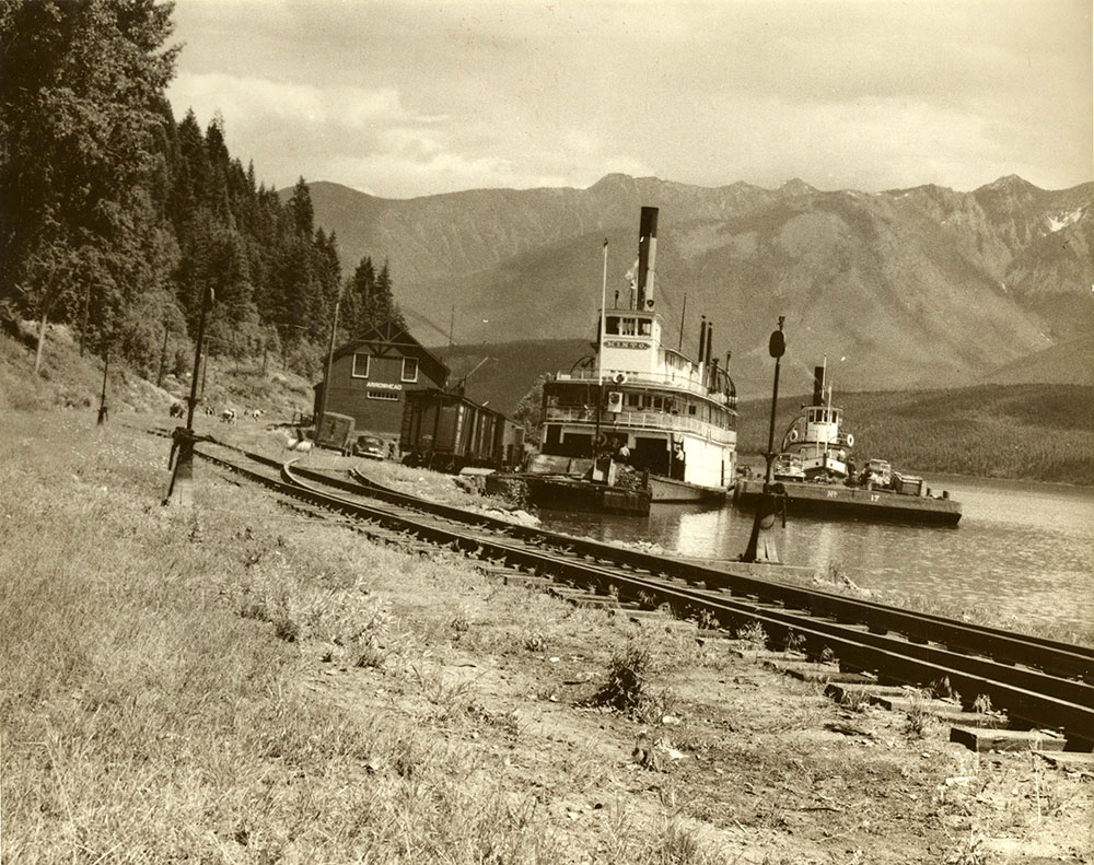 Sur cette photographie en noir et blanc, deux bateaux à vapeur sont amarrés sur le bord de la rivière, à côté d’une voie ferrée. Un petit bâtiment se trouve à la fin du chemin de fer, près des bateaux. On aperçoit des arbres à la gauche et des montagnes à l’arrière-plan.