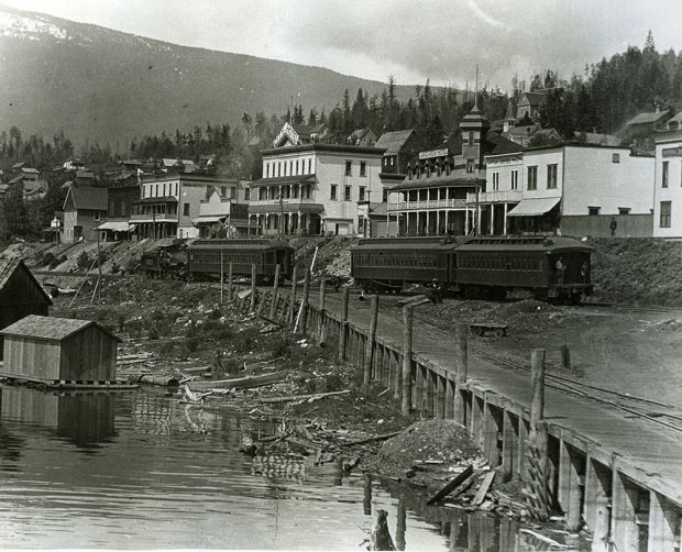 Cette photographie en noir et blanc montre une ville au bord de l’eau. La rue abrite des magasins et des maisons. Devant ces bâtiments, il y a une ligne de chemin de fer avec quelques wagons sur la voie. Un quai se trouve devant la voie ferrée. Une partie d’une montagne et des arbres se trouvent en arrière-plan.