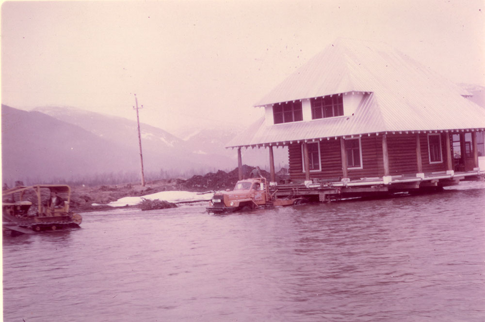 Une maison en bois rond de deux étages est déplacée de l'autre côté de la rivière sur une remorque à plate-forme de la compagnie Apex Moving Company. La remorque est dans la rivière, les roues presque couvertes. Un tracteur se trouve à gauche, avec un câble le reliant à la remorque de déménagement. Les montagnes sont en arrière-plan. 