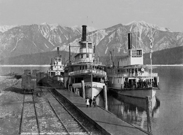 Photographie en noir et blanc. Trois bateaux à vapeur figurent côte à côte sur l’eau, à côté d’un quai. Des gens sont à bord des bateaux et quelques-uns sont sur le quai. Des voies ferrées se trouvent à côté du quai et il y a des montagnes à l’arrière-plan.