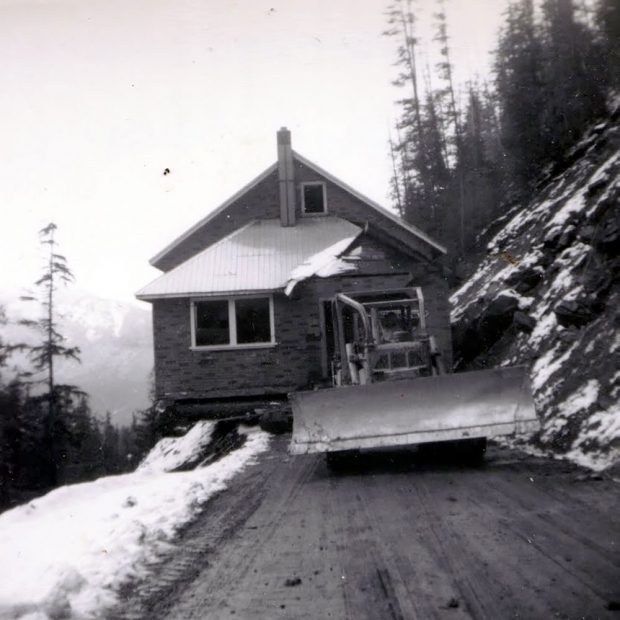 Photographie en noir et blanc d’un petit bâtiment en brique tiré par un chasse-neige sur un chemin de terre. Le bâtiment est légèrement suspendu sur le côté d’une falaise. Il y a de la neige au sol et des arbres de chaque côté de la route.