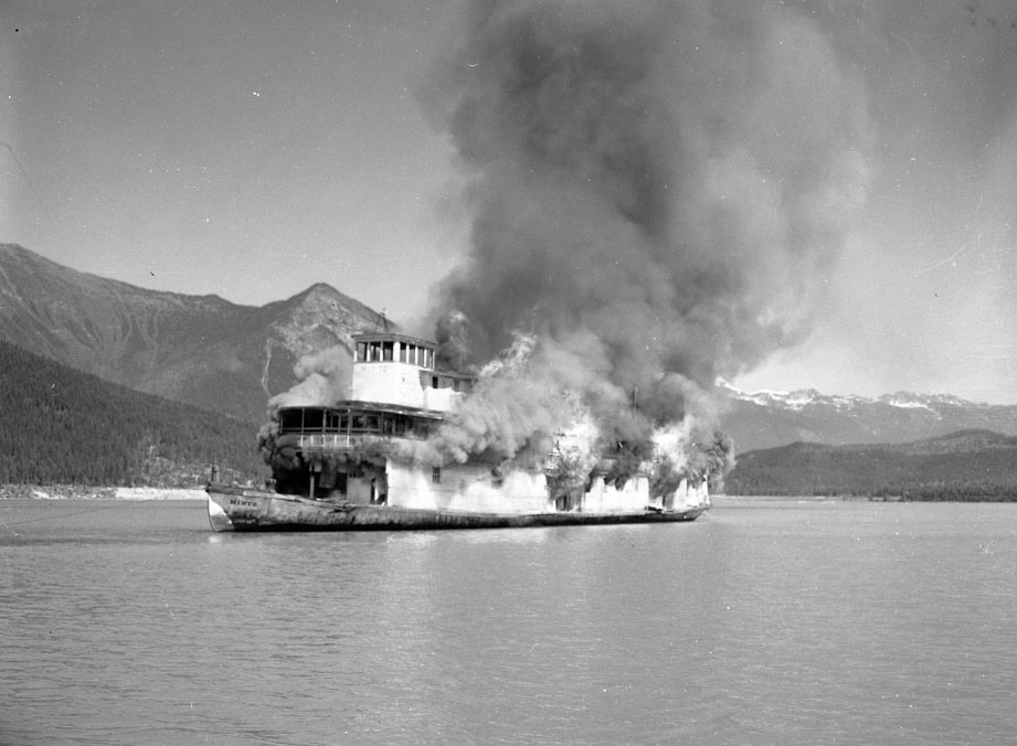 Photographie en noir et blanc d’un bateau à vapeur enflammé, au milieu d’un lac, duquel se dégage d’énormes nuages de fumée. Il y a des montagnes en arrière-plan.