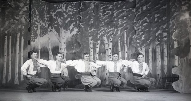 Photographie en noir et blanc de cinq danseurs sur une scène. Les danseurs sont accroupis sur leurs pieds et ont leurs bras sur les épaules des autres. L’arrière de la scène est une murale de peupliers faux-tremble.