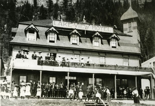 Photographie en noir et blanc d’un hôtel à deux étages qui porte l’enseigne « Lakeview Hotel » . Une foule de personnes se tiennent sur les deux balcons superposés de l’hôtel. Elles sont toutes habillées chic avec vestons-cravates, robes et chapeaux.